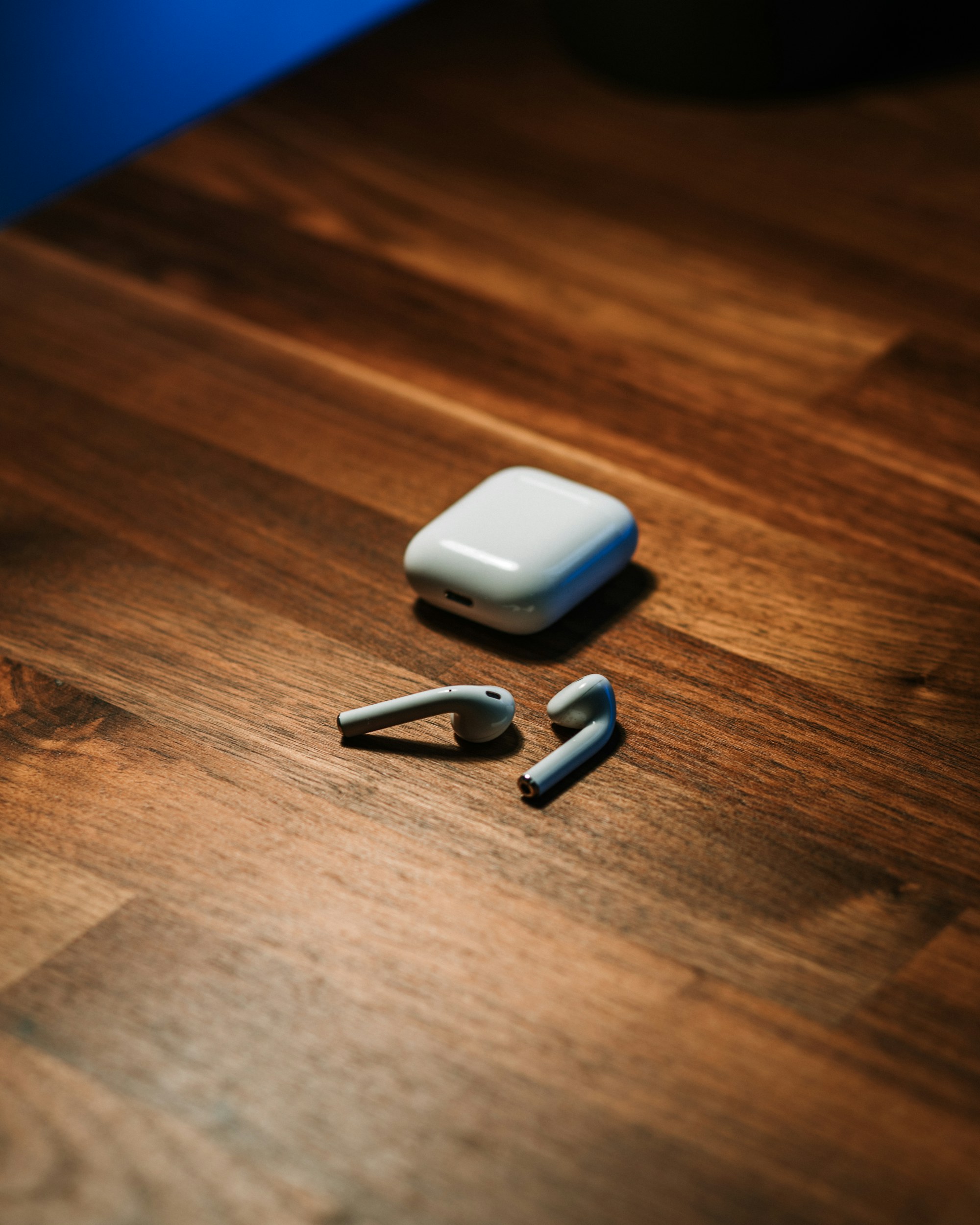 Earlier-generation Apple AirPods and charging case on a wooden table, used as an editorial photograph for AirPods 5 charging-case news.