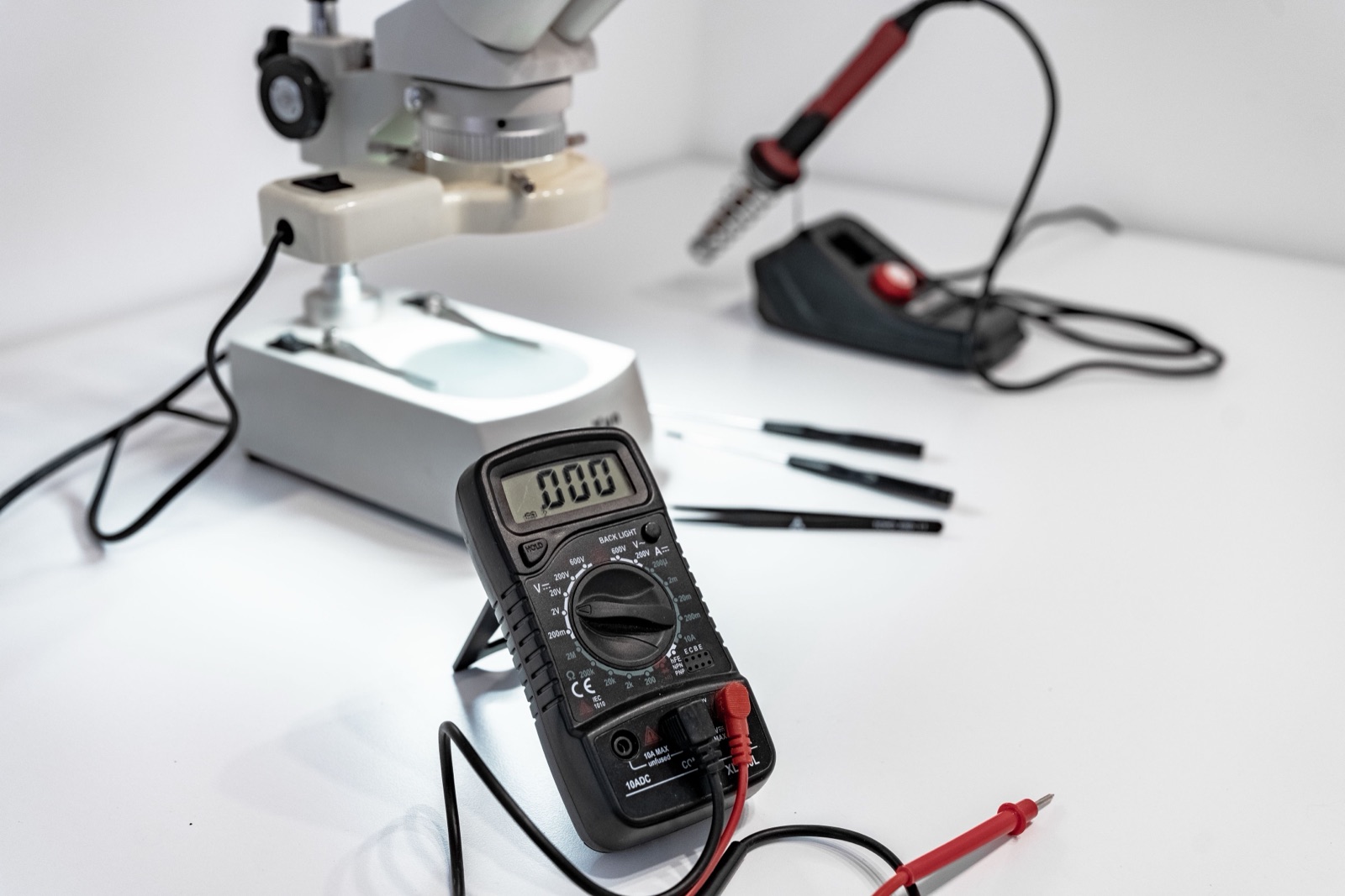 A digital multimeter on an electronics workbench, with a microscope and soldering tools behind it.