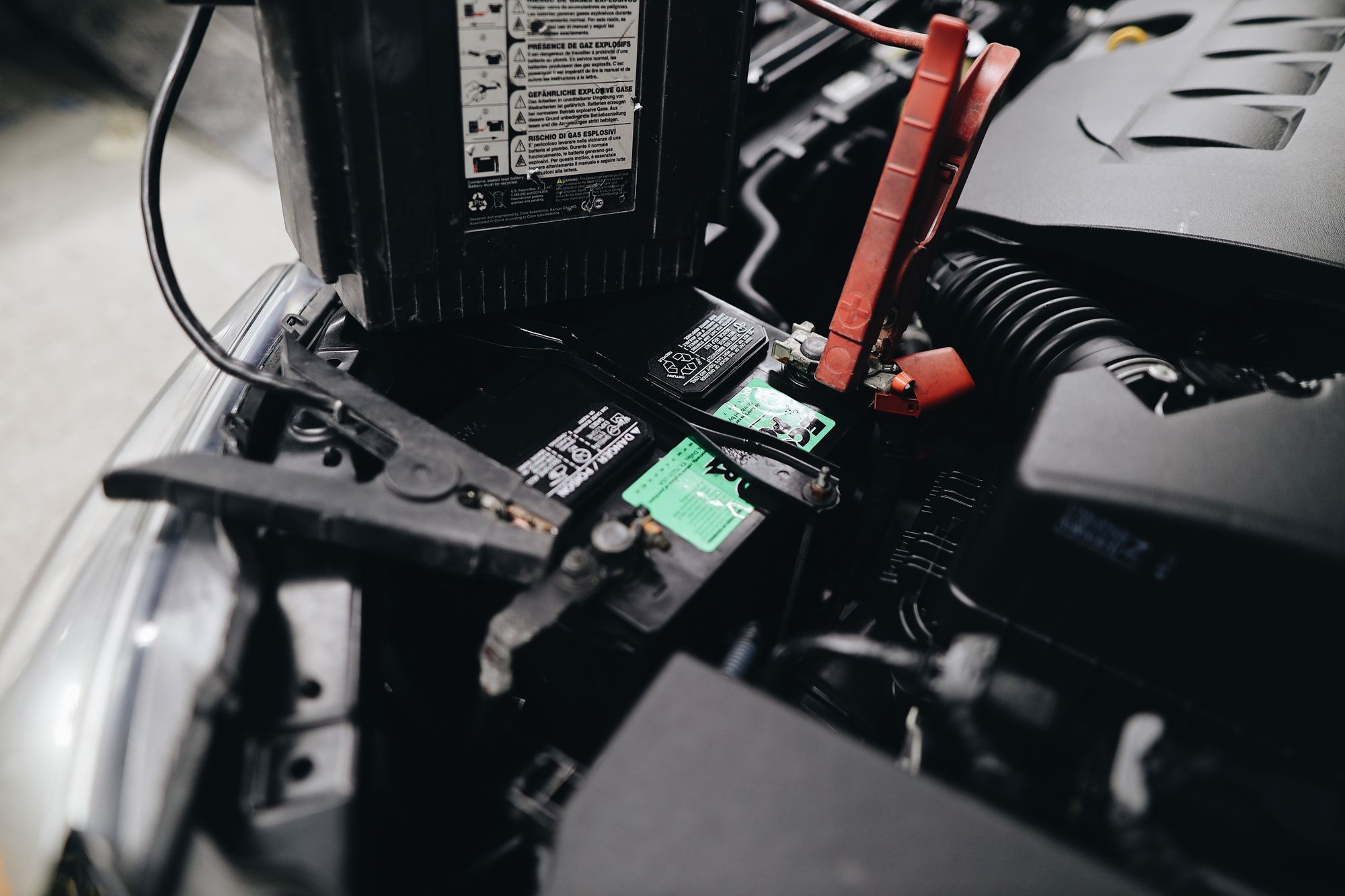 A real editorial photograph of a vehicle battery connected to charging or jump-start cables in an engine bay.
